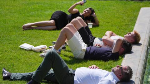 Four people are seen lying down on the grass in hot weather. A man in white shirt and green trousers is wearing sunglasses, then there are two people next to him - one shielding his eyes - lying down. At the back a woman is seen holding a phone up, either taking a phone call or a photograph.