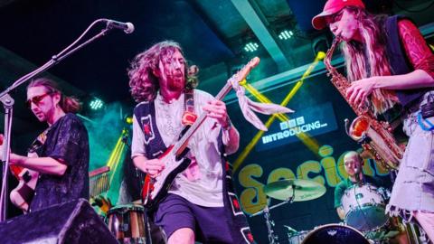 Four musicians playing instruments on a stage. Two at the front on the left are playing guitars in front of a mic. The one on the left has long hair and is playing a saxophone while a man in the back is playing the drums. They are performing at a music festival.
