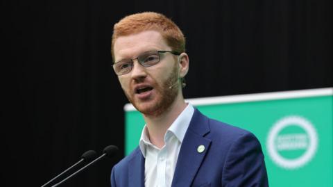Ross Greer at the Green Party manifesto launch standing in front of a huge green poster in a navy blue suit jacket and white shirt