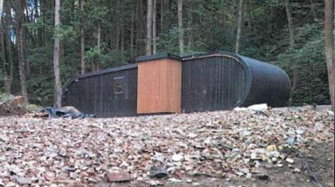 Just in front of many trees in a woodland, a black oval-shaped accommodation pod sits in a clearing. The floor is covered in bricks and rubble.