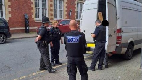Four police officers, three men and one woman are standing on the street next to a white van. There is a black circle over someone's head, who has been arrested. One of the back doors of the van are open.