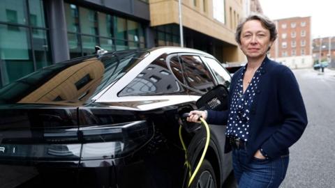 Ford UK's managing director Lisa Brankin wearing blue jeans, a navy blue blouse with white polka dots and a navy blue cardigan. She is standing next to a black Ford Capri connecting it to an electric charger and smiling at the camera