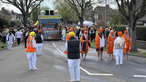 Image shows a residential road filled with hundreds of people, in the middle is a decorated lorry which the crowd is close to on either side. In the foreground of the image are a group of Sikh men dressed in traditional orange clothes and turbans. 