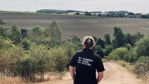 A woman is stood overlooking acres of fields and countryside with her back to the camera. She has dark hair pulled back and is wearing a black t-shirt which says "no farmers, no food, no future"