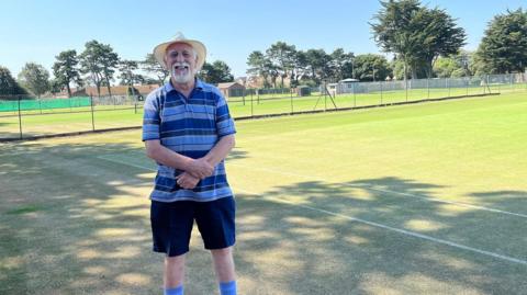 Organiser Chris Holt stands in front of a link fence inside of which are lots of tennis courts marked out with white lines and mesh nets. Chris wears a blue horizontally striped t-shirt, dark blue shorts and blue socks. It's a very sunny day so he has a sun hat on to shade his face. He has white hair and beard. 