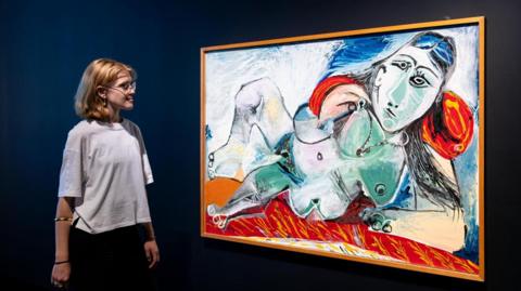 A woman with long blonde hair and glasses wearing a white T-shirt is looking at a Picasso displayed in front of her. It is a large canvas depicting a woman reclining and is painted in a surreal style. 