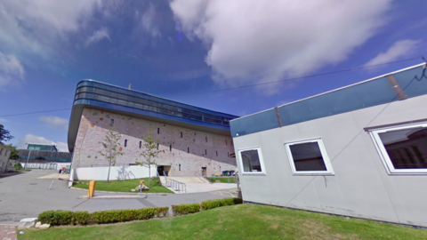 Penryn campus, University of Exeter. The screengrab has been taken looking up at the campus building which has been built with a light pink brick. There are narrow window slots across the building and a glass floor at the very top of the building.