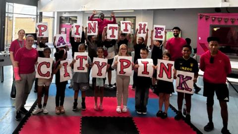 Team members from The Way wear bright pink T-shirts and hoodies, with children and young people holding up letters spelling out 'paint the city pink'. They are stood on a colourful pink, blue, grey and black mat.
