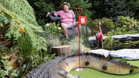 David Lawrence sits at the jungle themed hole with his arm around a model monkey, while models of an alligator tail and a flamingo are visible in the foreground