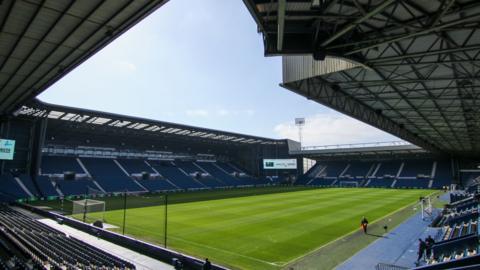 Inside The Hawthorns stadium where the green pitch and some dark blue seats are visible in front of blue sky