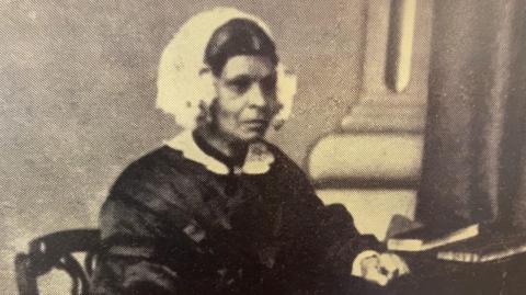 An old black and white photograph of Mary Ann Machem in Victorian clothing (black dress with long puffed sleeves) and white bonnet sitting at a desk, looking into the distance. She's resting one hand on the desk next to two books.