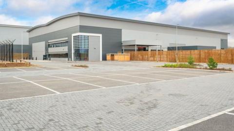 A grey building at a business park in the background, going from almost left to right in the photo. A wooden fence is to the side of it and in the foreground of the image are more than a dozen parking spaces.