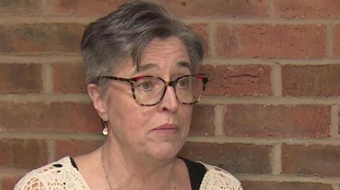 Gill Gray. She has short grey hair and is wearing large glasses. She is wearing a loosely knit beige cardigan.