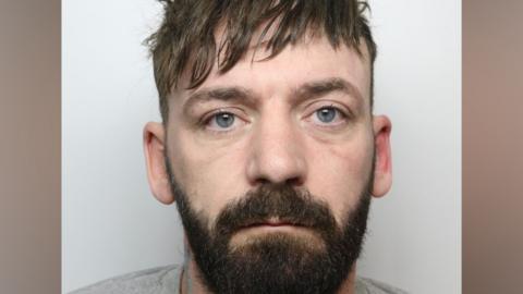 A police mugshot of a man with brown hair and beard wearing a grey jumper