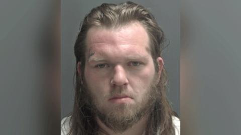 An expressionless man stares at the camera in a police custody photo. He has long brown hair and a beard, and an undecipherable written tattoo over his right eyebrow.