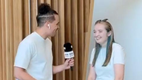 A BBC reporter with a bun talking to a girl with blue and blonde hair