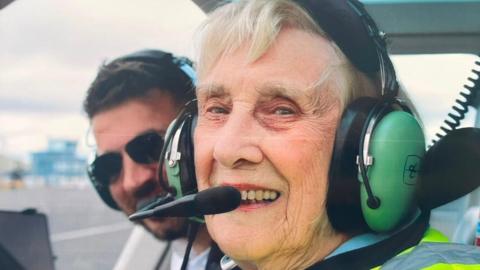 Joan Airey is sitting in the front seat of a helicopter and smiling brightly at the camera. She has short, white hair and is wearing a large green headset. A man is poking his head out from behind her.