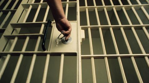 A prison cell door. The bars of the door are white. There is a hand putting a key in the lock of the door.