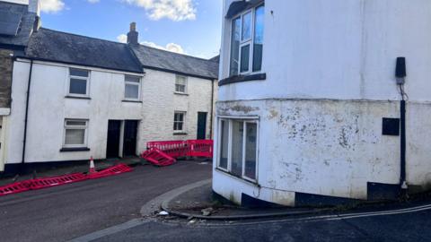 A white building on the corner of a road junction in Bodmin with masonry on the pavement after it broke off. Road barriers are in the background as part of a road closure set up. Some of the barriers have been tipped up on the floor.