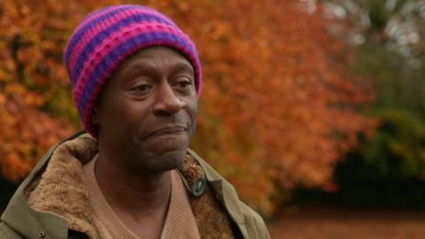 Mark Burnett stands in a park in autumn. He is wearing a purple and pink woolly hat and a green fur winter coat over a brown jumper.