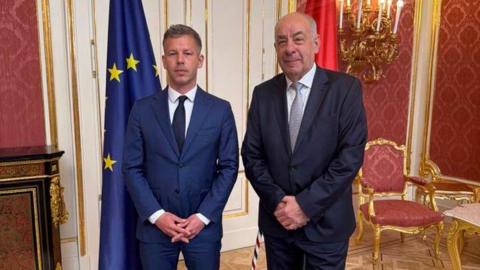 Péter Magyar stands beside President Tamás Sulyok. The two men wear suits and have their hands crossed in front of them.