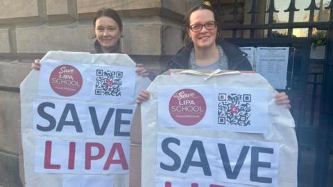 Two women with brown hair, one is wearing glasses, hold two banners up which say 'Save LIPA school' in blue and red large letters