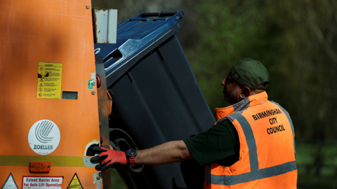 A refuse worker in an orange high-vis waistcoat and red gloves unloads a bin into the back of an orange bin lorry in April