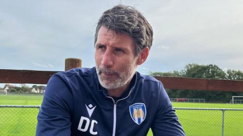 Danny Cowley, who has medium length, greying hair. He is sat in front of a football pitch while wearing a navy tracksuit top, which has the Colchester United crest on it. He is looking to the left of the camera with a slight frown.