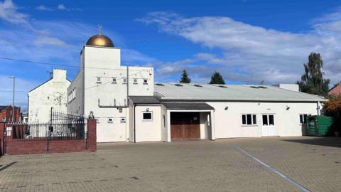A view of the outside of Northampton Central Mosque on a bright but cloudy day. The mosque is shown from its car park and is mostly white with a gold qubba at its highest point.