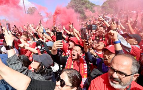 Liverpool fans celebrate winning their 20th league title. There are thousands of fans wearing red shirts and waving scarves. Red smoke is pouring out of flares held up in the air
