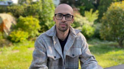 Myles is wearing a light grey jacket with a black t-shirt underneath. He is wearing black framed glasses and has a bald head with stubbly facial hair.