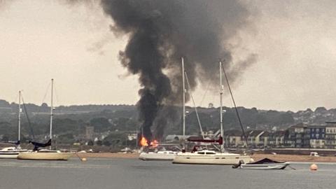 Thick black smoke pours goes into the sky from a large fire in Exmouth. Boats are on the water in front of the fire. Buildings are in the background.