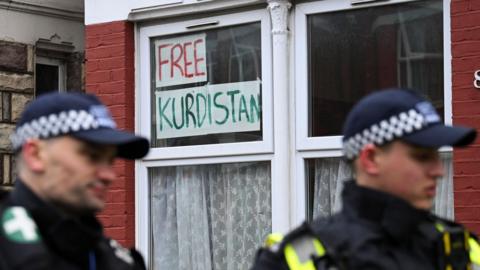 Two police officers in uniform standing in front of a residential window with a sign reading "Free Kurdistan" displayed, set against a red brick house with white-framed windows.