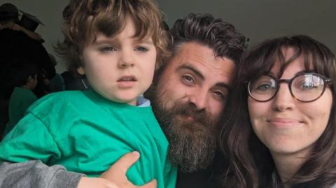 A child wearing a green shirt, and a man with a beard and woman with glasses are smiling in shot