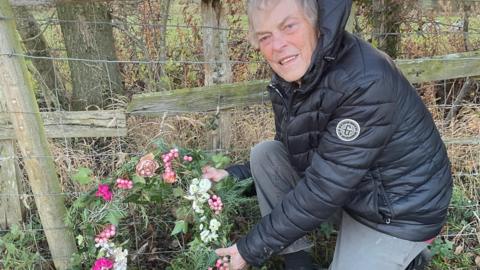 A man in a black puffa jacket and grey joggers lays a floral wreath next to a wooden and wire fence in a grassy field.