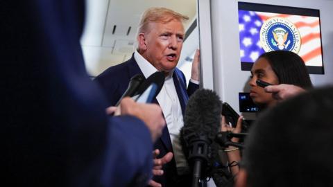 President Trump on Air Force One being interviewed by journalists. He is standing beside a video screen.