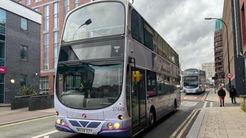 A First-branded purple double-decker bus travels along a road in Sheffield city centre, followed by another in the background.