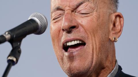 Bruce Sprungsteen sings into a microphone. He has grey hair and very thin grey stubble, with a gold earring in his left ear