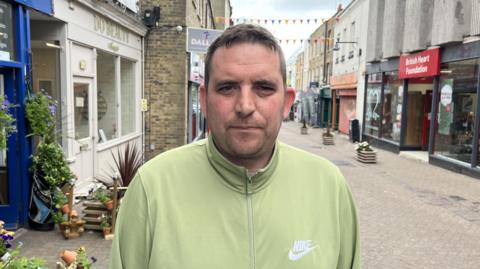 Dan Todd in a green zip-up jumper, looking at the camera. a high street is visible behind him.