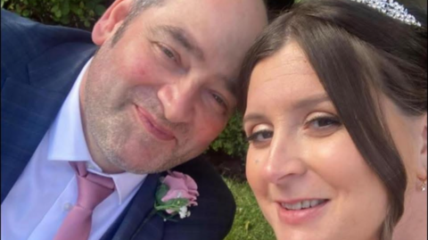 A selfie of Caroline and Robert on their wedding day. They are both smiling at the camera. Robert is wearing a dark blue suit with a white shirt, pink tie and pink flower button hole. Caroline is wearing a tiara and a white dress.