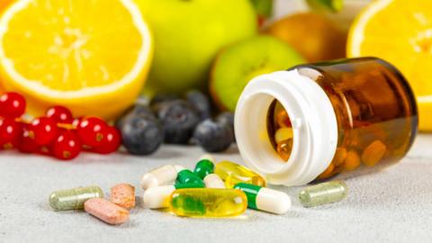 A bottle of pills on its side with a pile of vitamin pills next to it, in front of a row of fruit