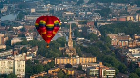 A multi coloured hot air balloon flying over Bristol. The city landscape, consisting of buildings and water, can be seen below. 