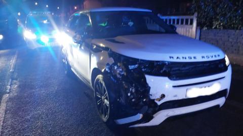A banged up white Range Rover. Front-right bumper and bodywork around the wheel has come away. There is a police car behind the Range Rover with it's blue lights on.