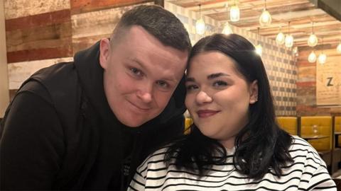 Ryan Storey has short black hair and is wearing a black top. He is standing next to his fiancée Anais who has black shoulder-length hair and is wearing a white and blue striped top