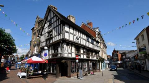 The corner of a two-storey black and white timbered building with pedestrianised streets running down both sides and market stalls with red and white awnings on the left hand side. There is bunting hanging over both streets.