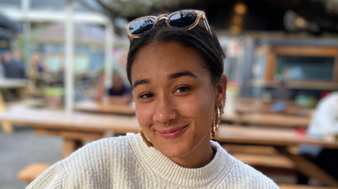 Beth, wearing a white top and with sunglasses perched on her head. She is sitting with some benches behind her which are out of focus