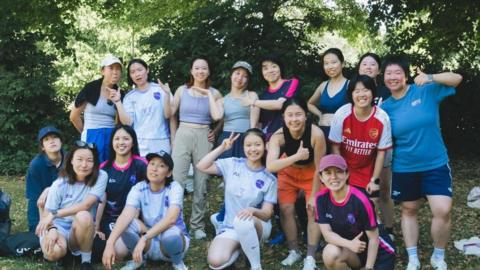 A group photo of the Screamer Alliance women’s football team, showing 16 players smiling and posing together outdoors under the shade of trees. Some are wearing football kits, including white and purple shirts and an Arsenal jersey, while others are in casual sportswear. Several players are crouching at the front while others stand behind them, making peace signs and thumbs-up gestures.