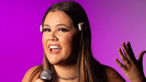 Ms Giorgio-Jones with long dark hair holding a microphone in her right hand and putting her left hand level with her face. She is in front of a purple background.