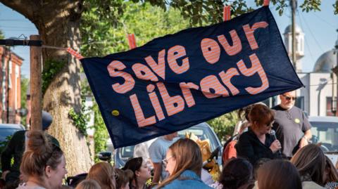 People standing below a banner saying Save Our Library
