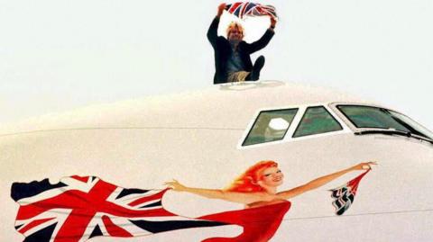 Richard Branson sits on top of a large white plane - only part of the plane can be seen. The windows for the pilots cockpit and underneath where there is the painting of the Scarlet Lady. She is smiling, looking like she is flying forward, wearing a read leotard and holding a billowing union jack flag behind her.